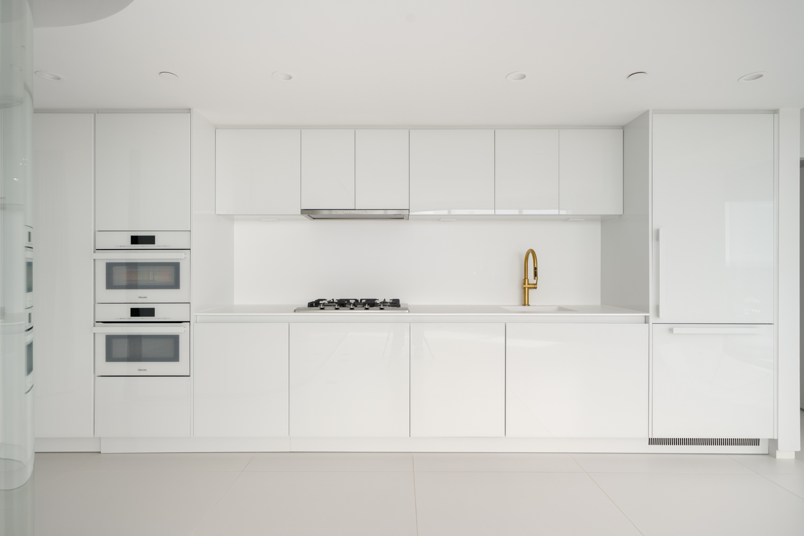 Minimalist modern white kitchen with built-in oven, stove, gold faucet, and sleek cabinets in a clean, bright setting.
