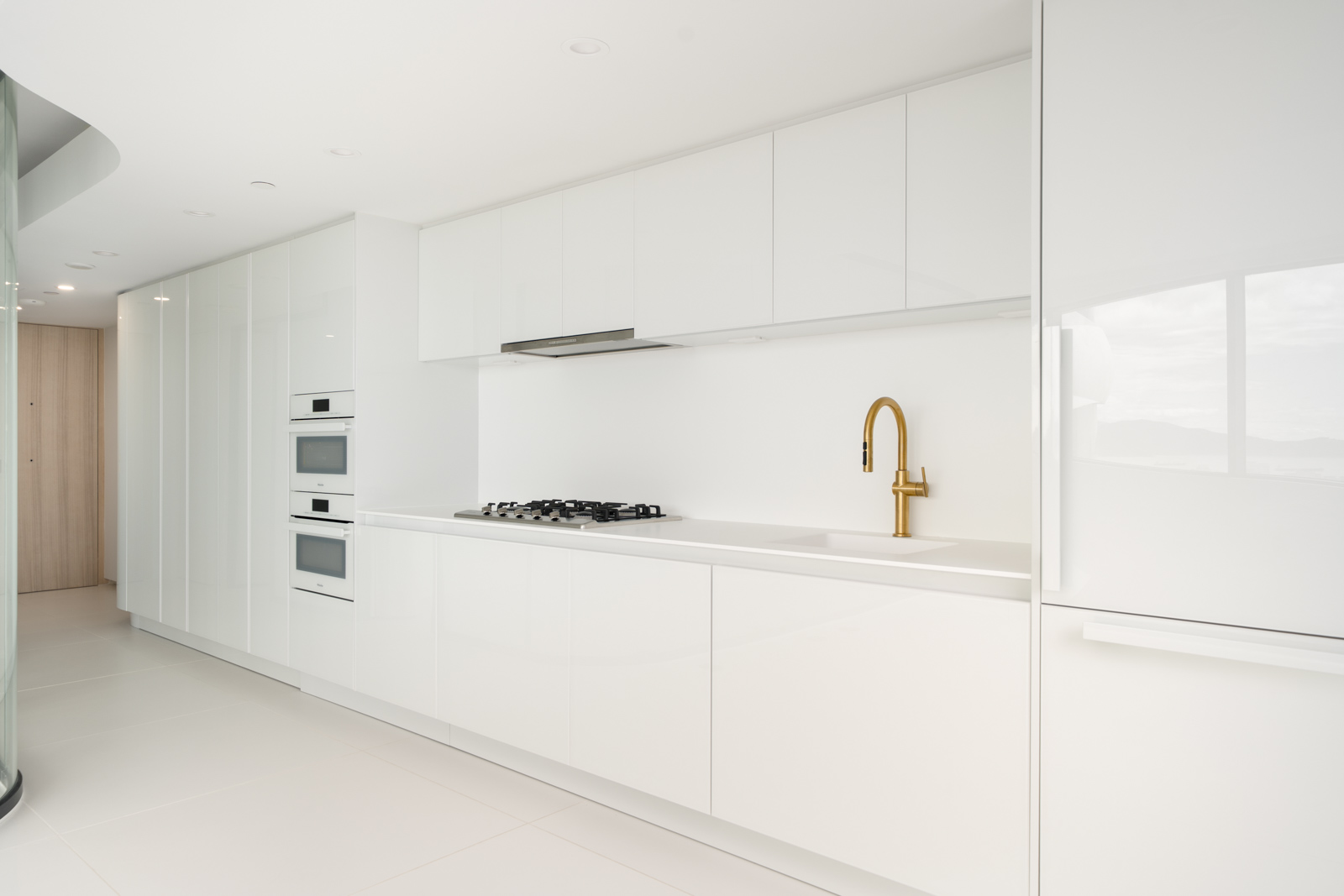 Modern white kitchen with built-in appliances, minimalist cabinets, a gas cooktop, and a gold faucet on a white countertop.