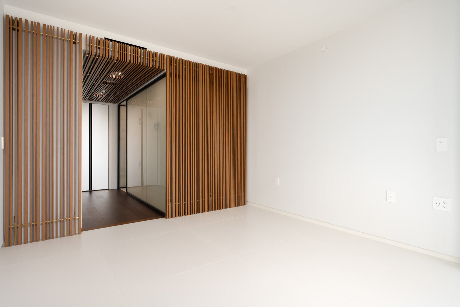 Minimalist, empty room with white tile floor, white walls, and a wooden slat partition leading to a hallway with wood flooring and glass walls.