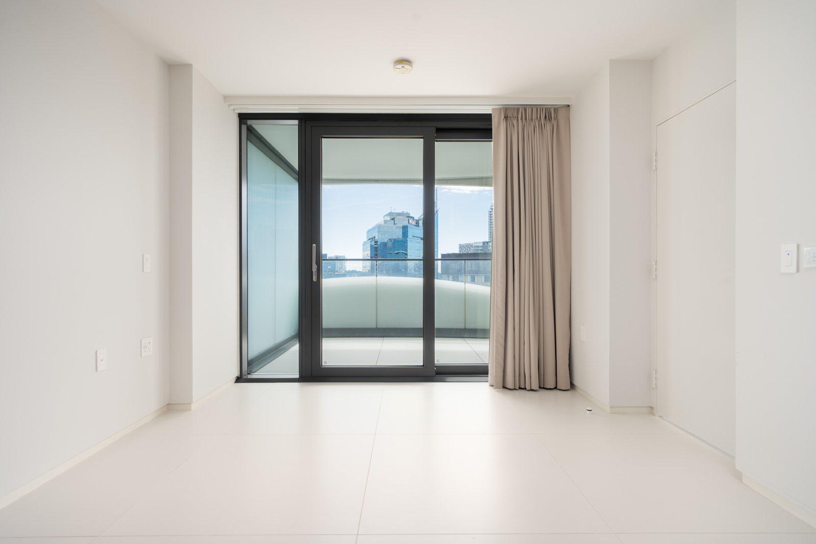 Empty modern room with large glass doors leading to a balcony, city buildings visible outside, neutral walls and floor, beige curtains on one side.