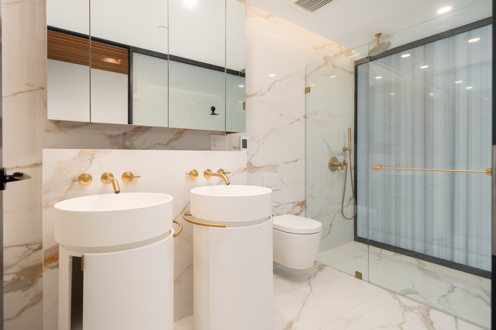 Modern bathroom with marble walls and floor, two white pedestal sinks, gold fixtures, a wall-mounted toilet, and a glass-enclosed shower.
