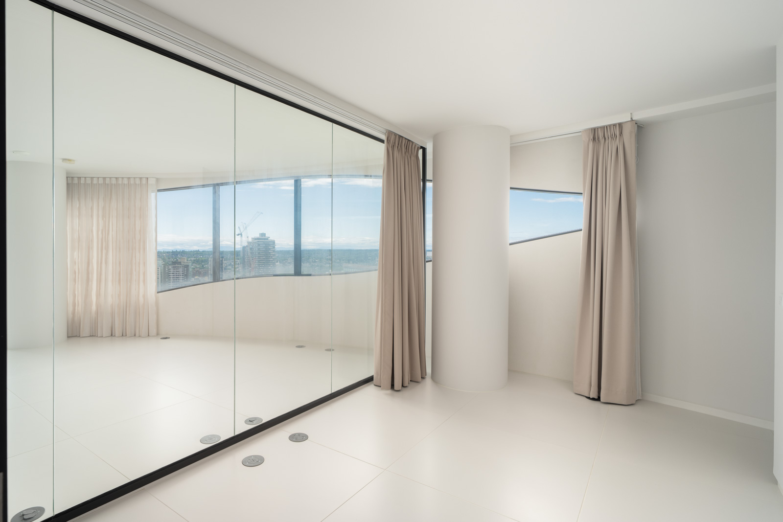Minimalist, modern room with large floor-to-ceiling windows, beige curtains, white tile floor, and mirrored sliding closet doors reflecting the cityscape outside.