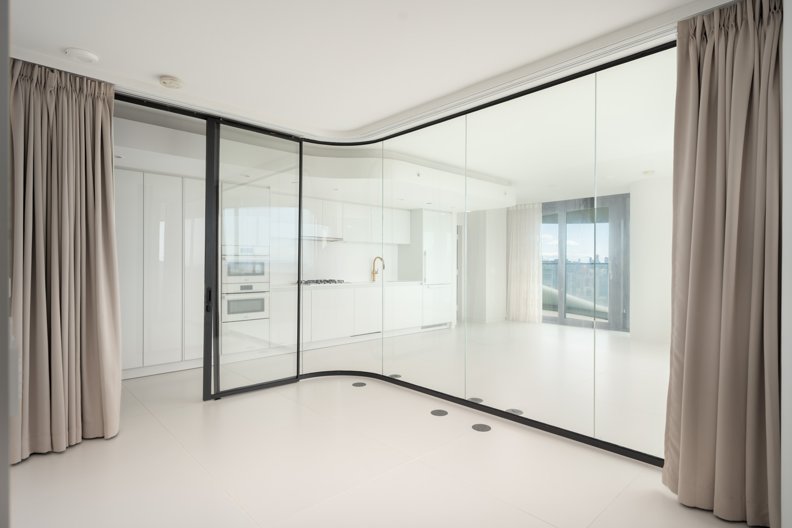 Modern, unfurnished apartment interior with floor-to-ceiling glass walls, beige curtains, white kitchen cabinets, and a large window offering a city view.