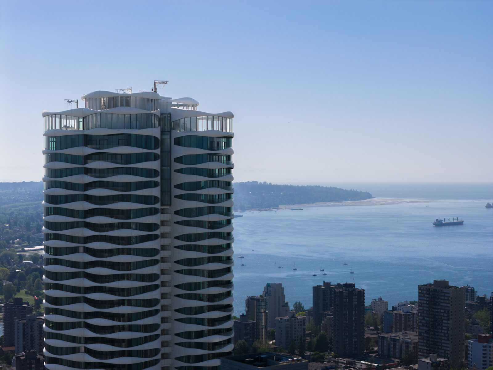 A modern high-rise building with wavy white balconies stands in front of a coastline with ships and a distant shoreline under a clear blue sky.