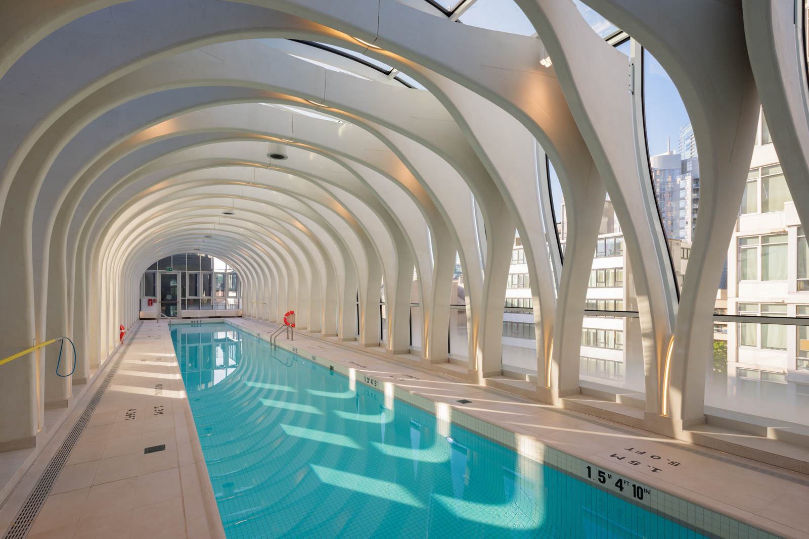 Indoor swimming pool with arched ceiling and large windows, surrounded by modern buildings; natural light illuminates the space.