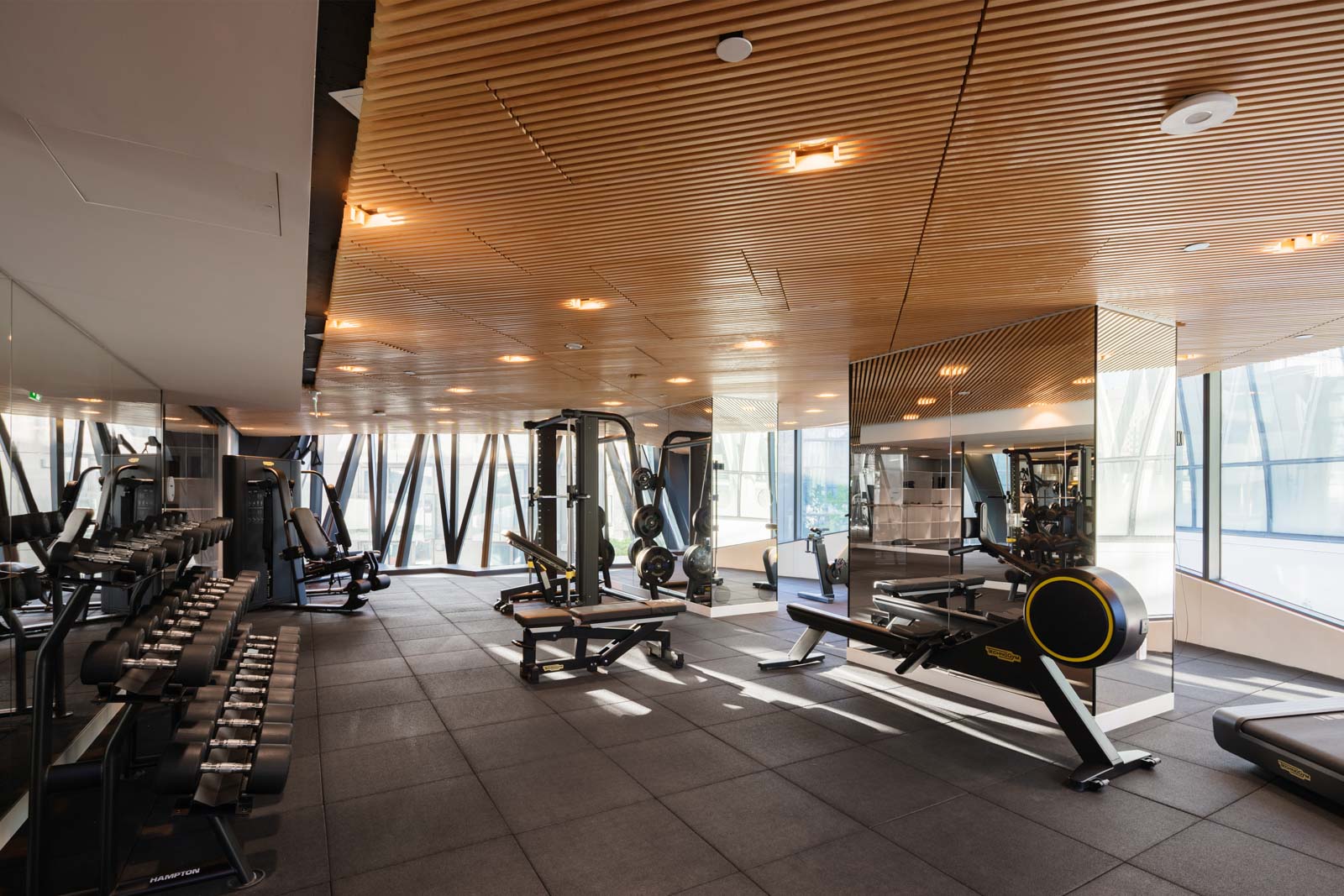 Modern gym with large windows, weight racks, exercise machines, rowing machine, and wood-paneled ceiling. The space is well-lit with natural and artificial light.