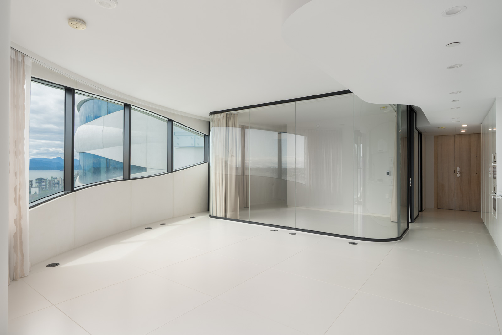 Spacious, modern apartment interior with large curved windows, white floors and walls, and a glass-walled room in the corner.