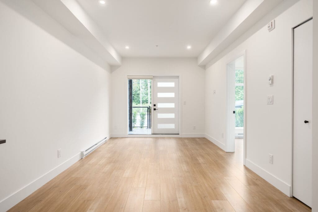 Empty, modern room with light wood flooring, white walls, recessed lighting, and a white front door with horizontal glass panels leading to a balcony.