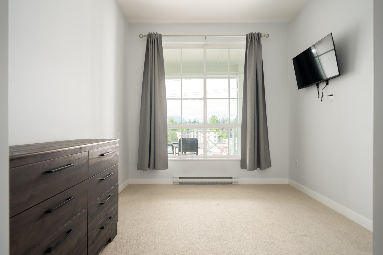 A small, empty bedroom with beige carpet, a wooden dresser on the left, a wall-mounted TV on the right, and a window with gray curtains overlooking a balcony.