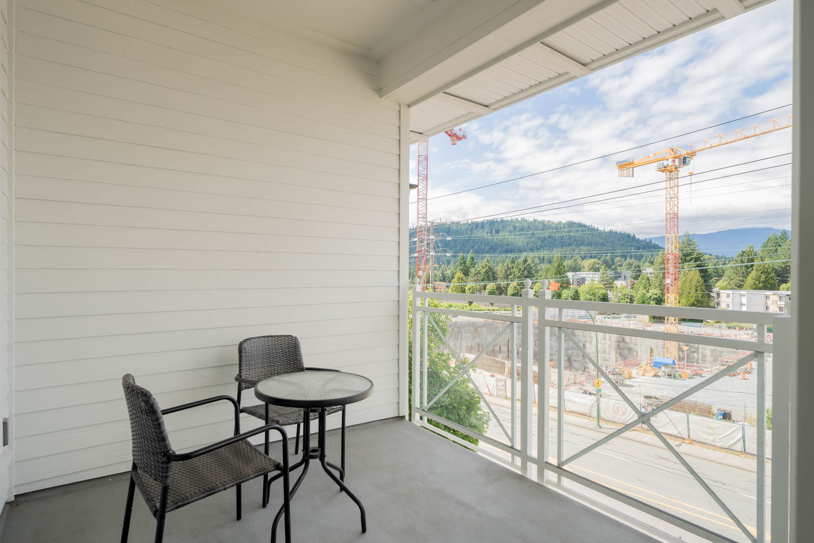 A small balcony with two chairs and a table overlooks construction cranes, a forested hill, and a partially cloudy sky.