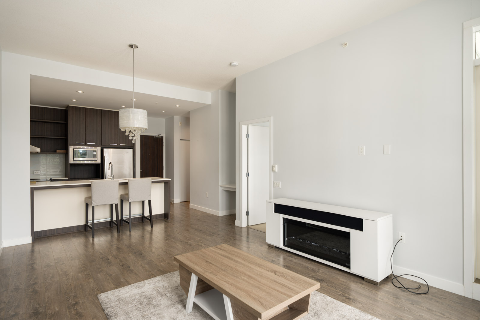 Modern, unfurnished apartment with open kitchen, bar seating, stainless steel appliances, wood flooring, and a minimal living area with a coffee table and entertainment unit.