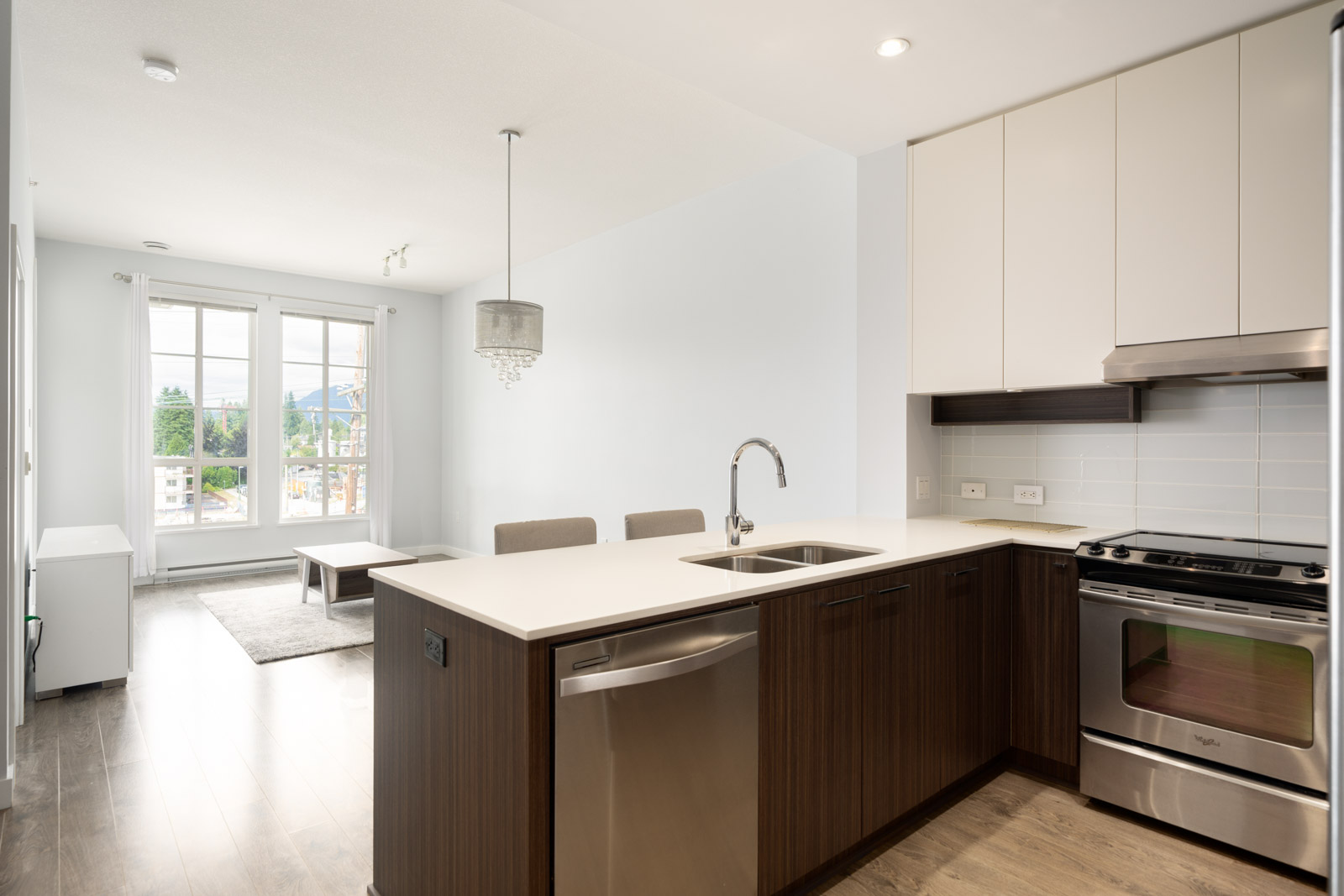 Modern kitchen with stainless steel appliances, a large island with a sink, and an open layout leading to a bright living area with large windows and a small dining table.