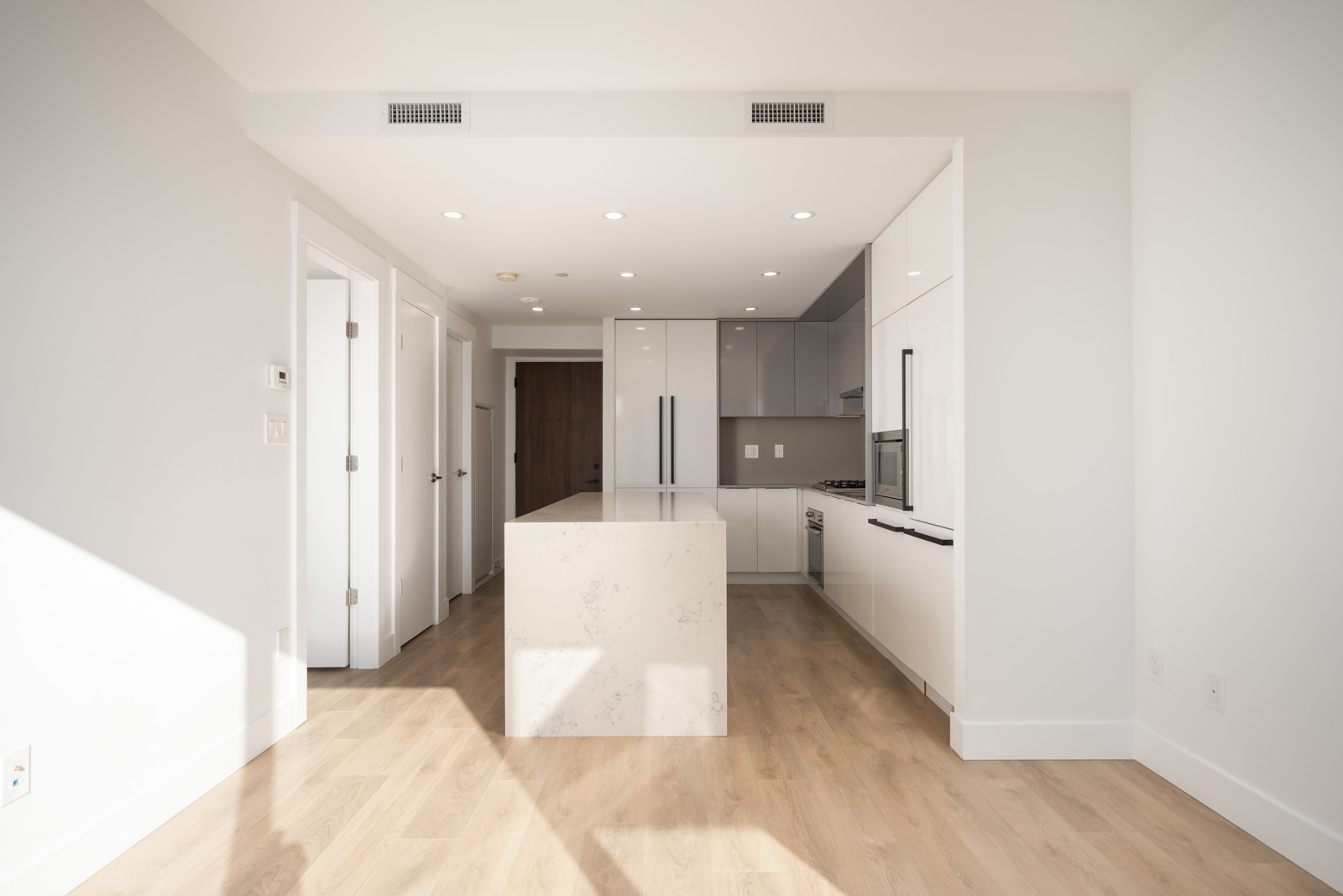 Modern, unfurnished kitchen with a central island, white cabinets, light wood flooring, and recessed lighting. Doors to other rooms are visible in the background.
