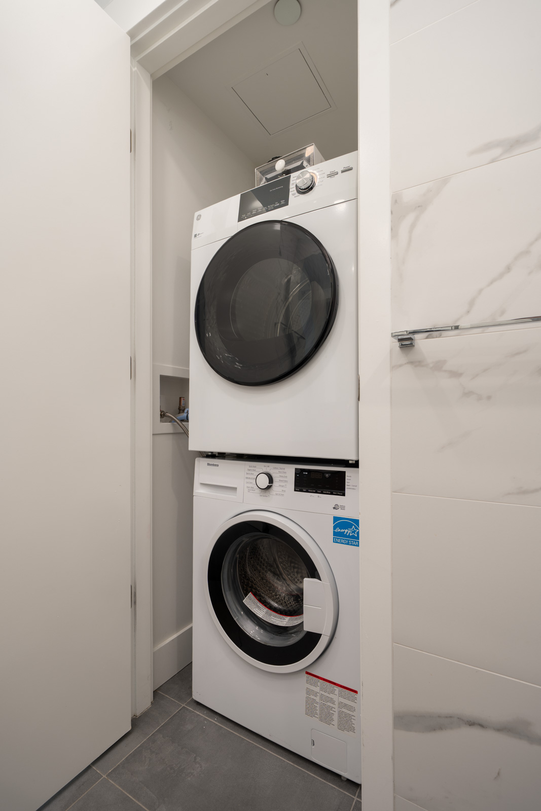 A modern, stacked washer and dryer set is installed in a small laundry closet with gray tile flooring and white walls.