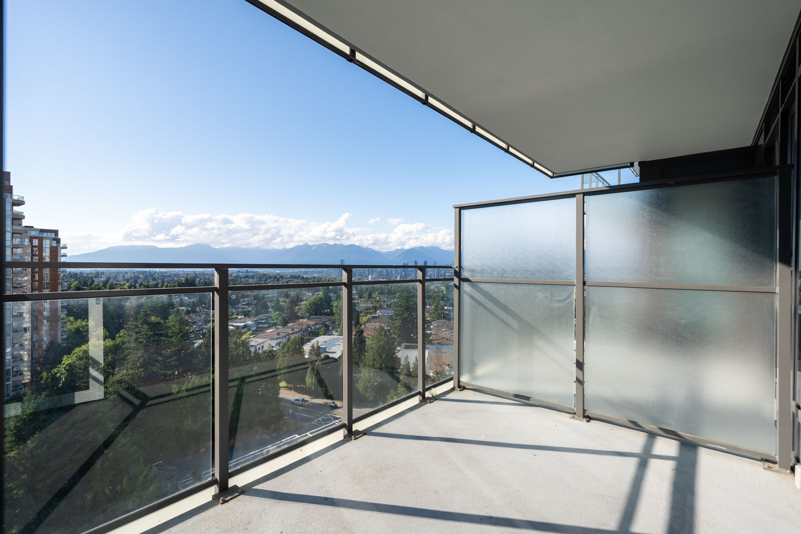 A modern apartment balcony with clear glass railings and frosted privacy panels, overlooking a cityscape and distant mountains under a blue sky.