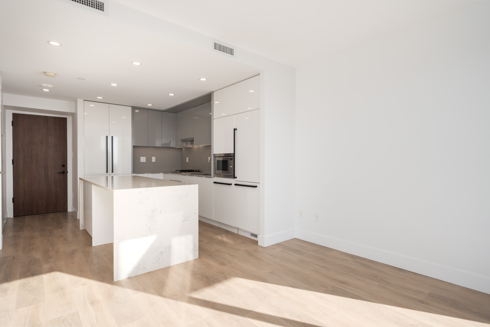 Modern kitchen with white cabinets, marble island, built-in appliances, wood floor, and minimal natural light entering from the right.