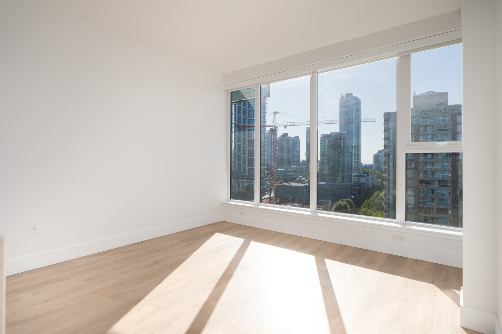 Empty room with light wood floors, white walls, and large floor-to-ceiling windows overlooking a cityscape with high-rise buildings.