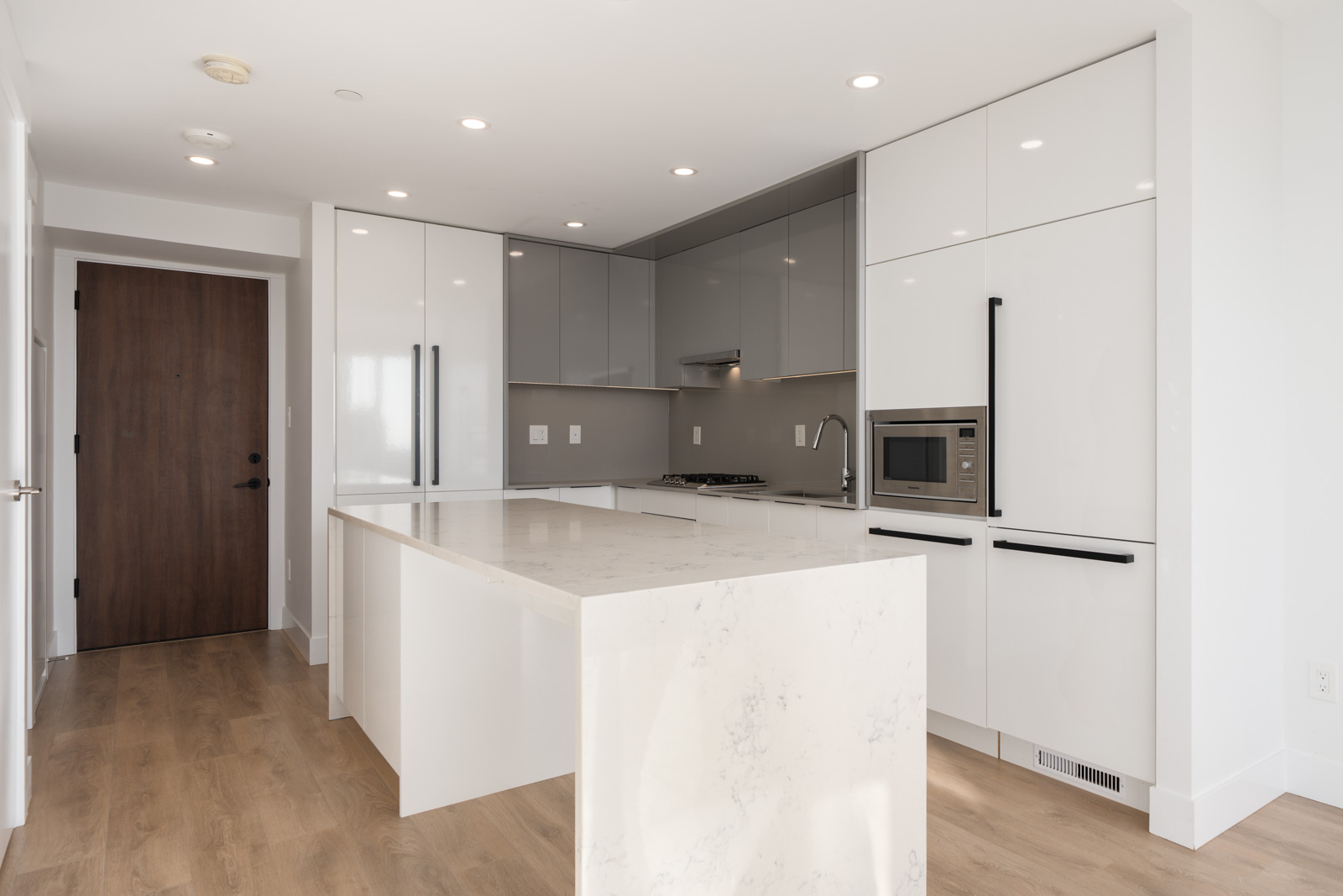 Modern kitchen with white cabinetry, gray backsplash, built-in appliances, large marble island, wood flooring, and recessed lighting.