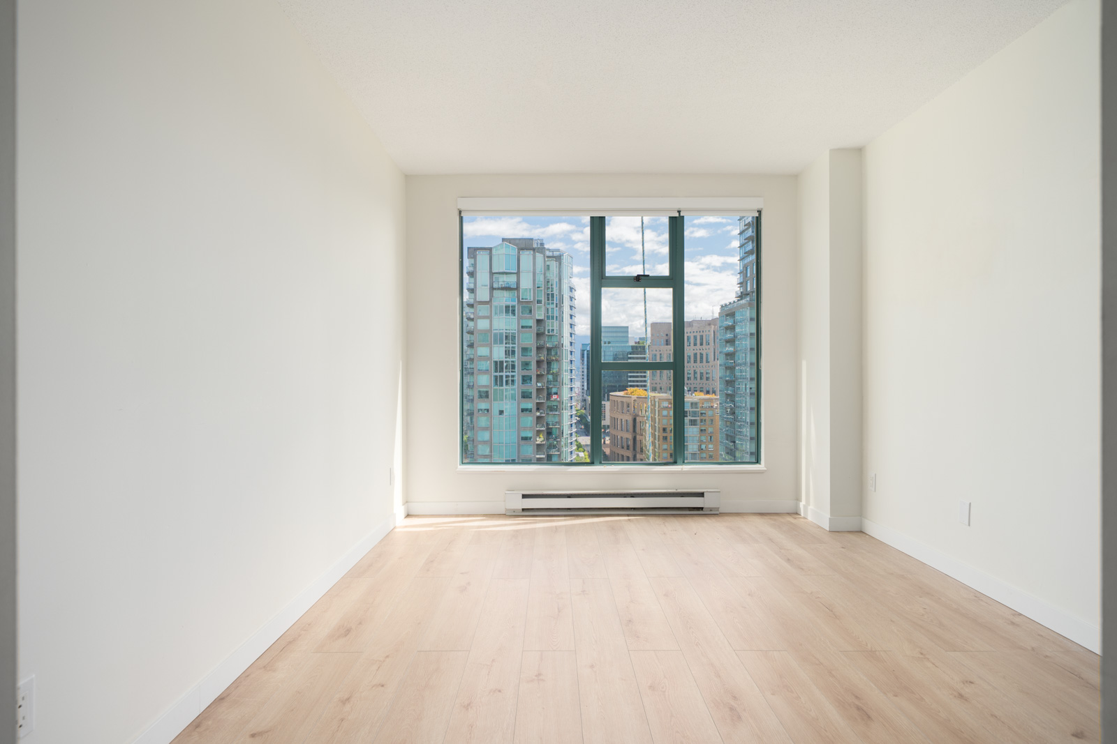 Empty room with light wood flooring, white walls, and a large window overlooking city buildings and a partly cloudy sky.