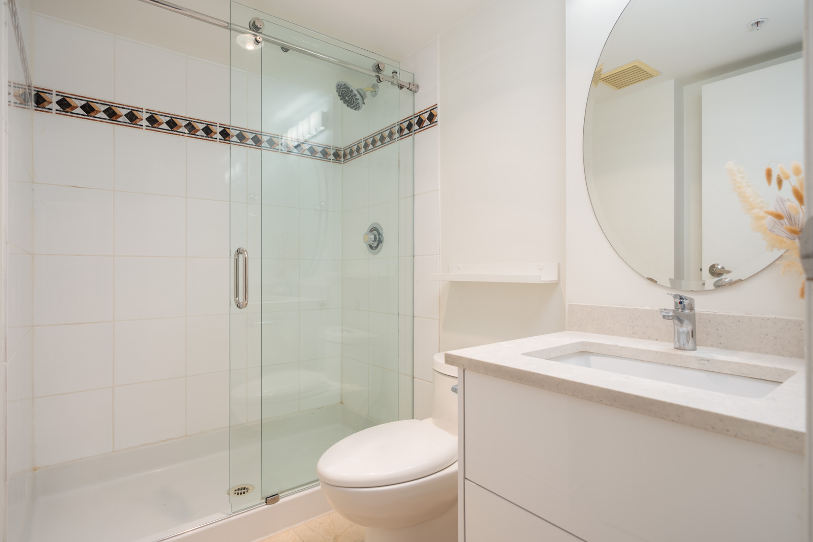 Modern bathroom with a glass-enclosed shower, white tiles, toilet, round mirror, and white countertop sink with a small vase of dried flowers.