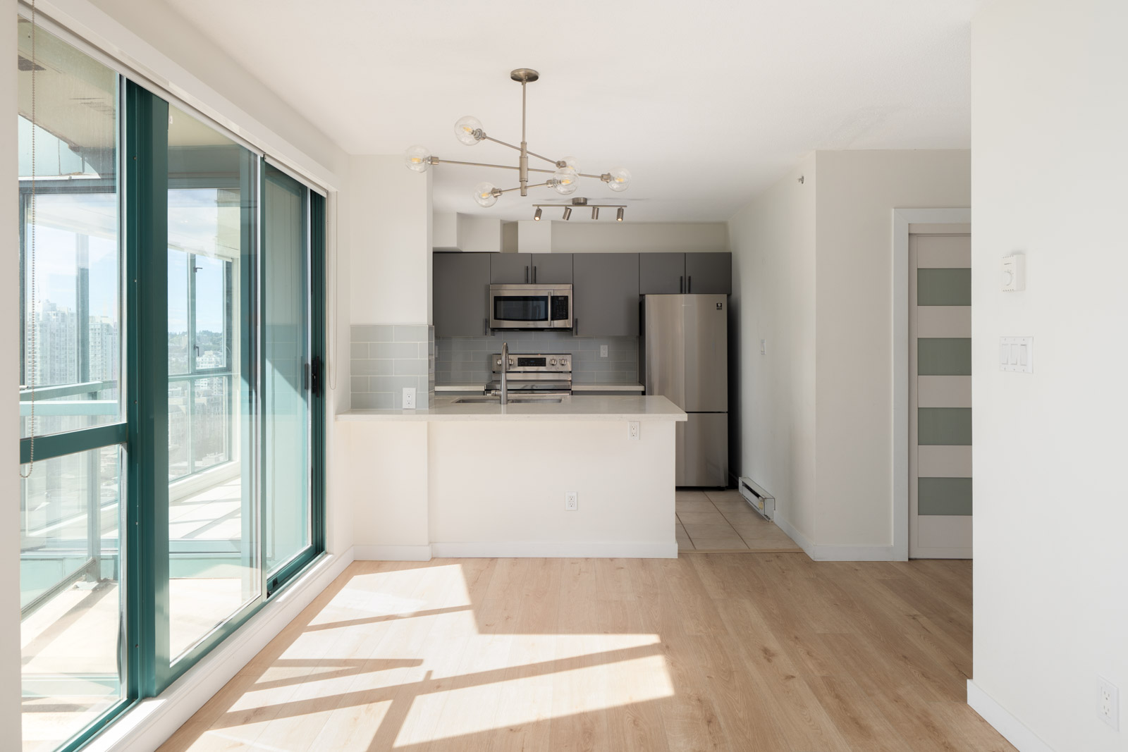Modern, unfurnished kitchen with stainless steel appliances, grey cabinets, light wood floors, and large windows letting in natural light.