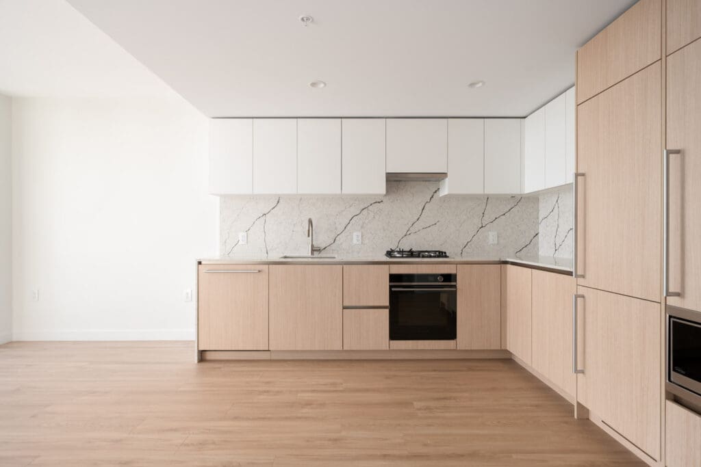 Modern kitchen with light wood cabinets, white upper cabinets, marble-patterned backsplash, built-in oven, stovetop, sink, and hardwood flooring.