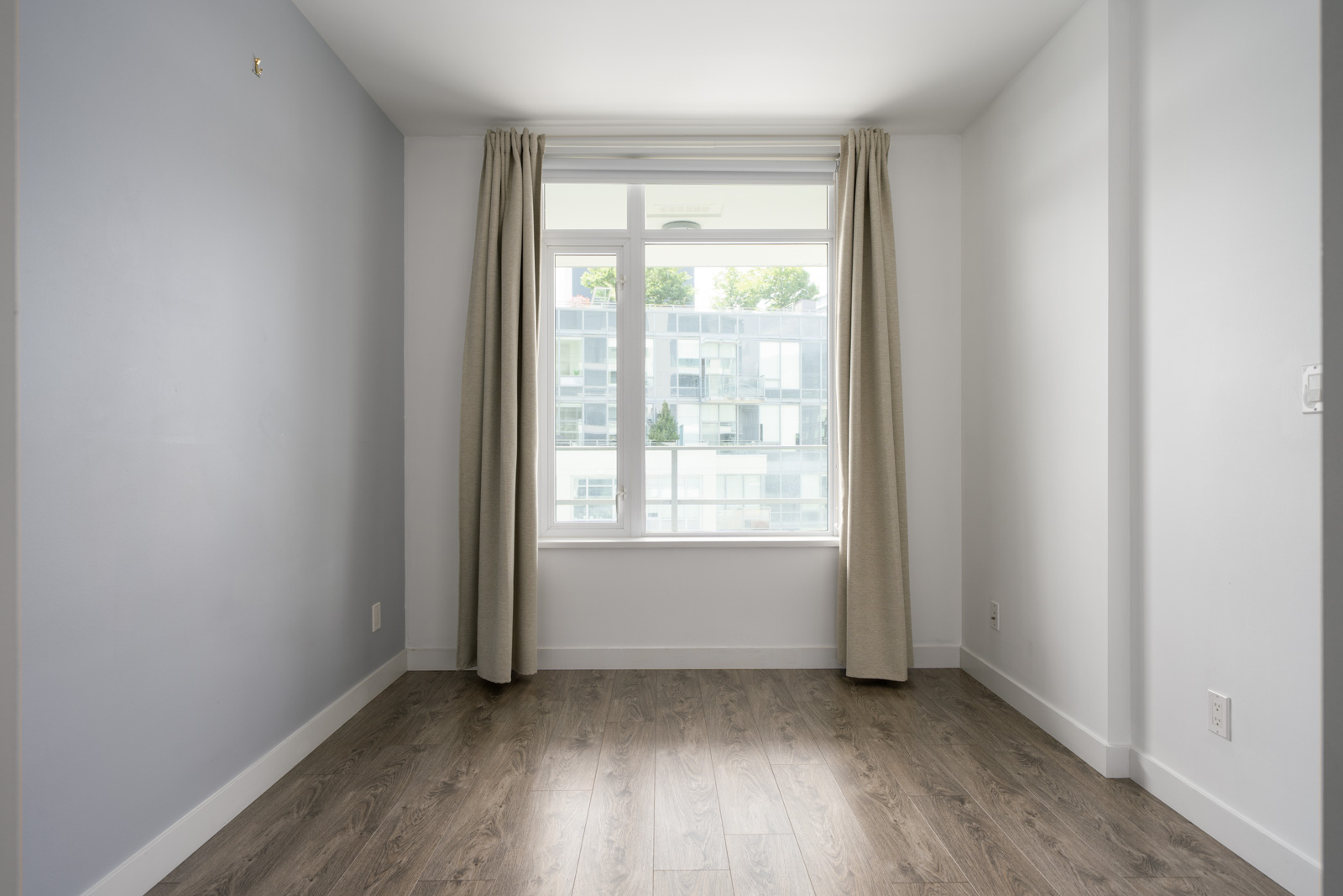 Empty room with light gray walls, wood flooring, beige curtains, and a large window facing an apartment building.