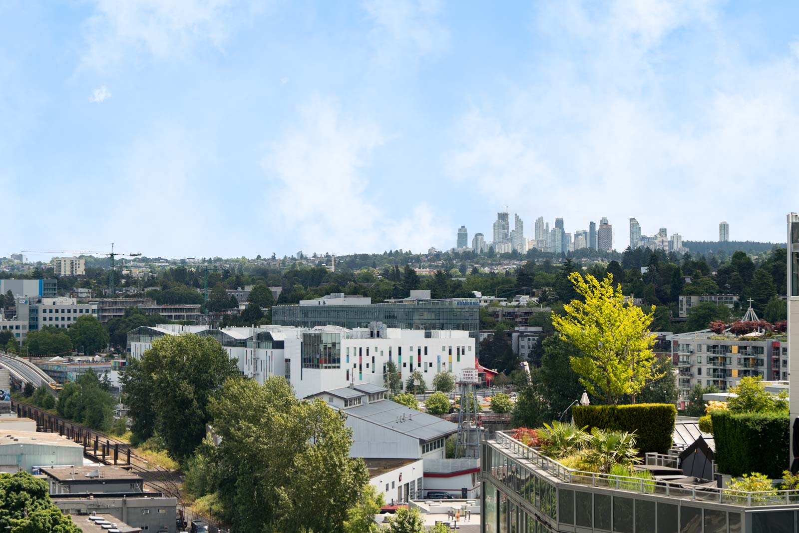 Cityscape view with a mix of modern and older buildings, trees, and distant high-rises under a blue sky with light clouds.