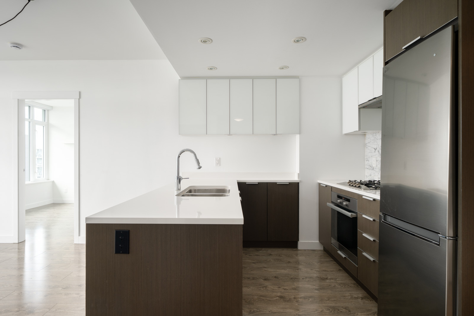 Modern kitchen with white countertops, wood cabinets, stainless steel appliances, a built-in oven, and an island sink; adjacent room visible through open doorway.