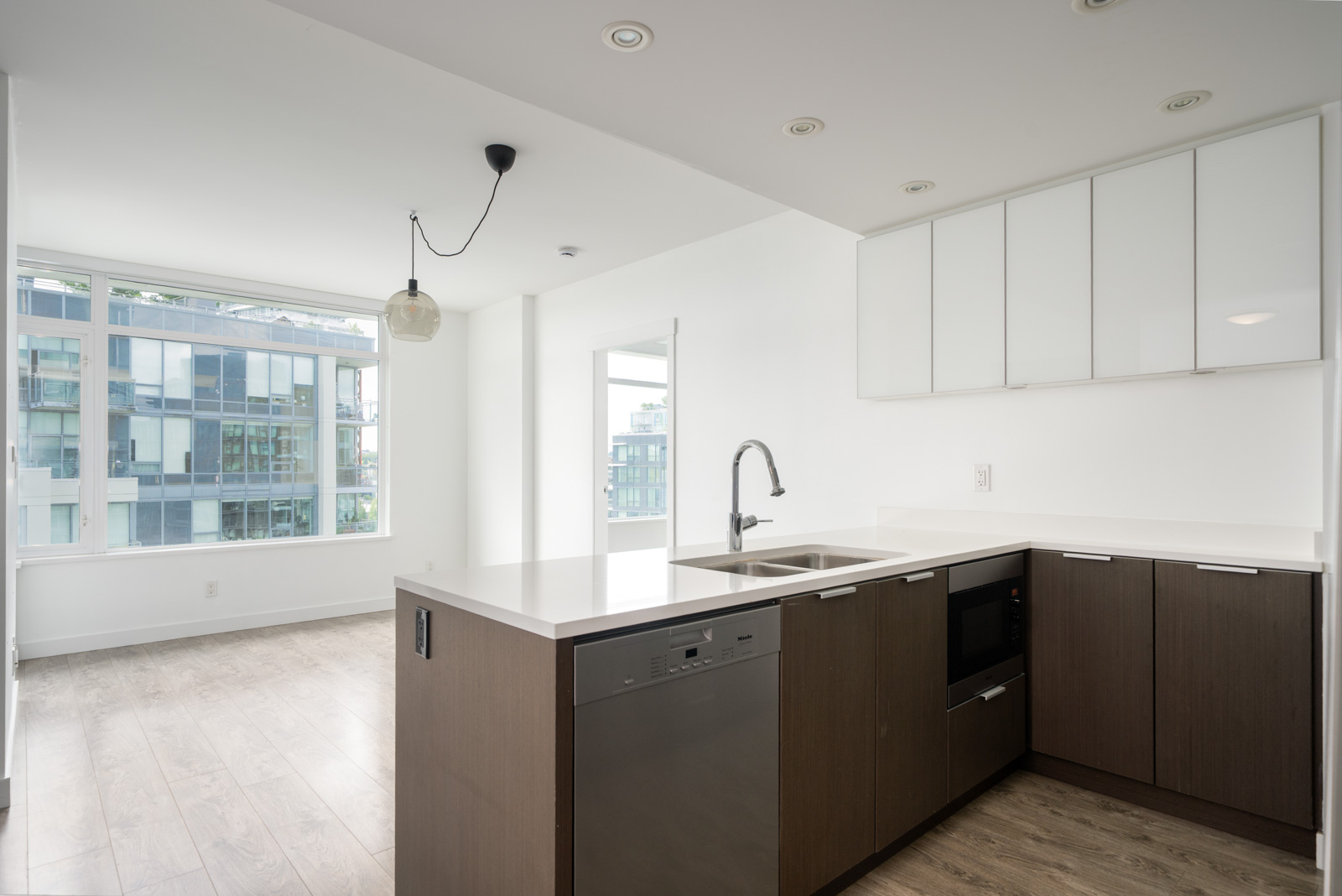 Modern apartment kitchen with white countertops, built-in appliances, and large windows allowing natural light into an open living area.