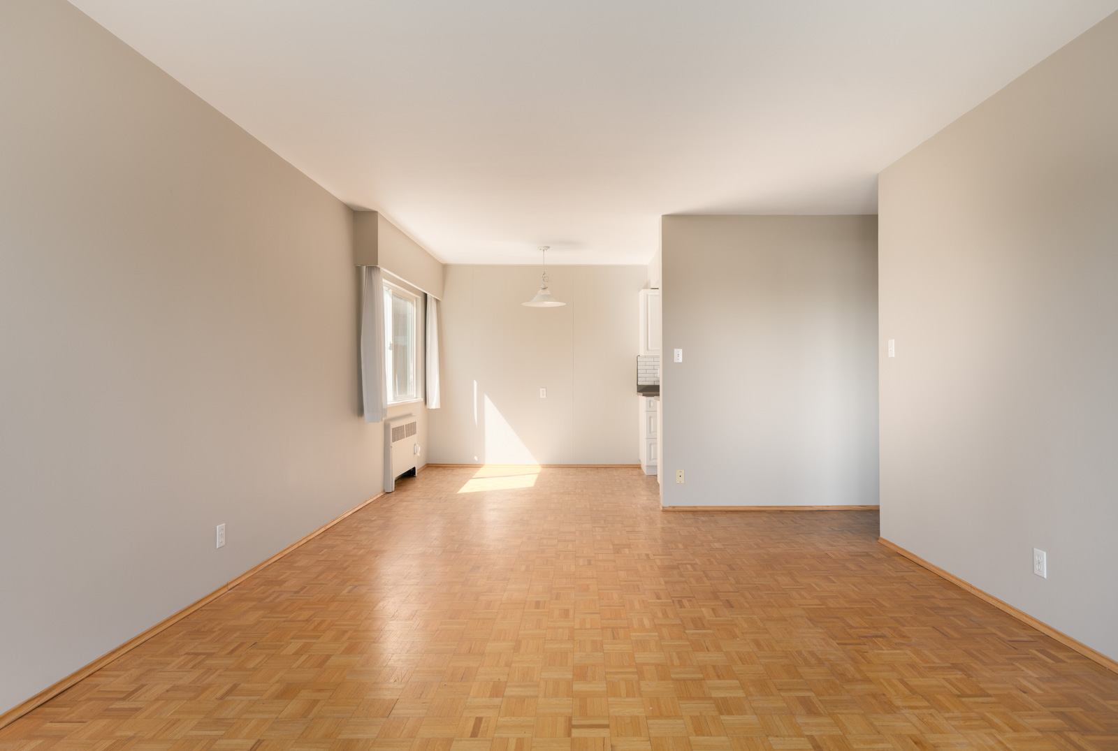 Empty room with light-colored walls, large window, parquet wood flooring, and a dining area with a hanging light fixture leading into a small kitchen space.