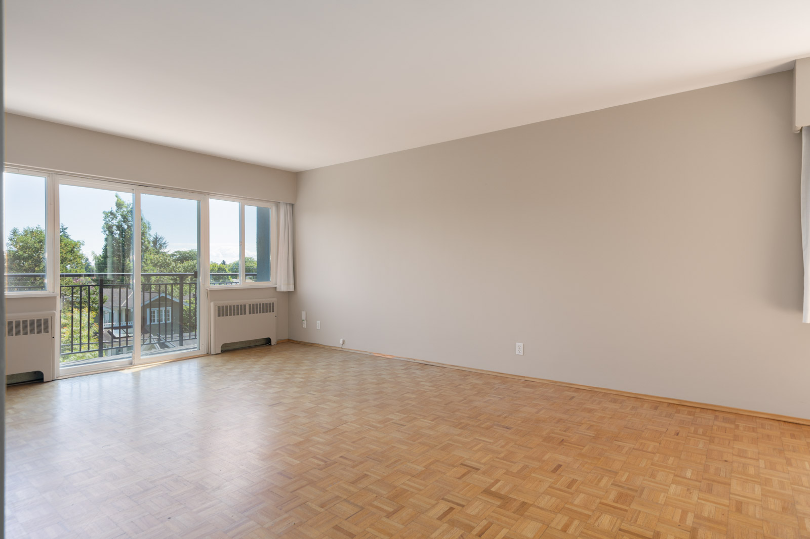 Empty room with parquet flooring, large window, and glass door leading to a balcony with a view of trees and greenery. Walls are painted light beige.