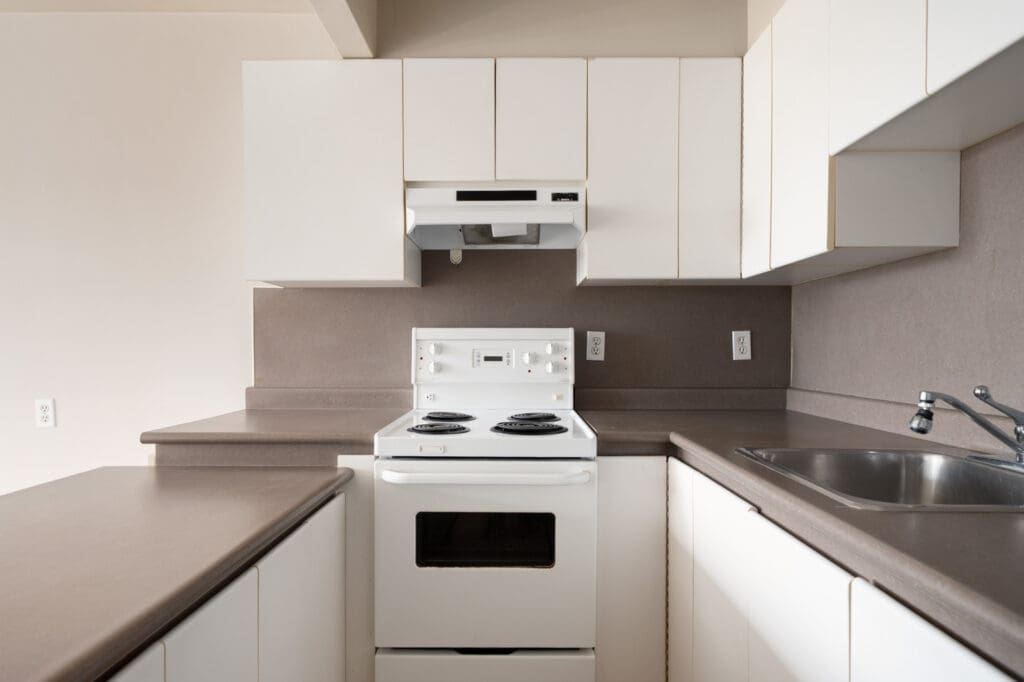 A small kitchen with white cabinets, a white electric stove, vent hood, brown countertops, and a stainless steel sink.